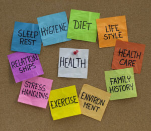 Image of health concept - word cloud or circle of contributing factors (diet, lifestyle, healtcare, family history, environment, exercise, stress, relationships, sleep, rest, hygiene), colorful sticky notes on cork bulletin board